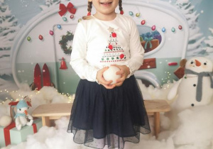 Zuzia trzyma w rękach białą kulę i pozuje do zdjęcia wśród zimowo- świątecznej scenerii.